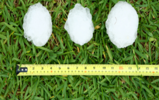 Three large, baseball-sized hailstones on green grass next to a yellow tape measure showing a diameter of over 3 inches, illustrating severe storm and hail damage in North Judson, Indiana.